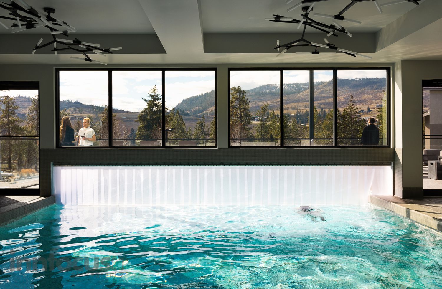 Indoor hotel pool with mountain views through large windows, featuring a waterfall feature and scenic forest backdrop.