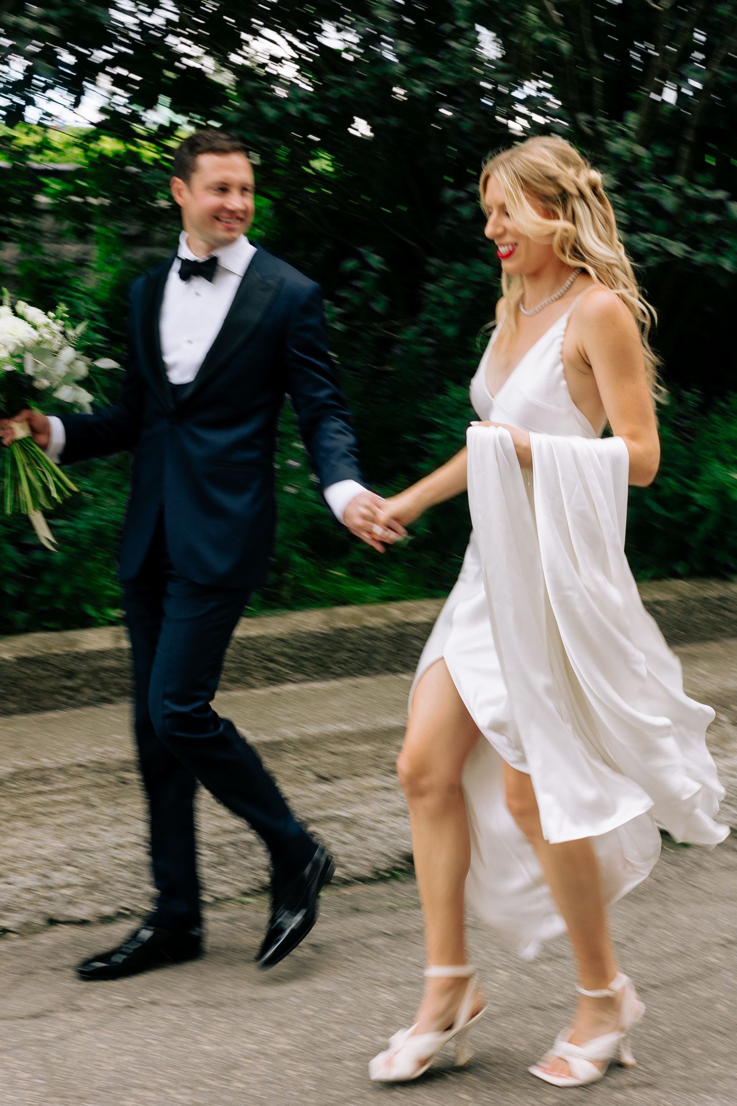 A newlywed couple walks hand in hand on a garden path, with the bride wearing a flowing white dress.