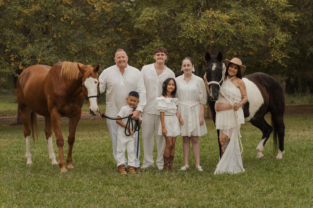 Family in white outfits posing outdoors with two horses on green grass with trees in background. Family photoshoot with a horse in sydney