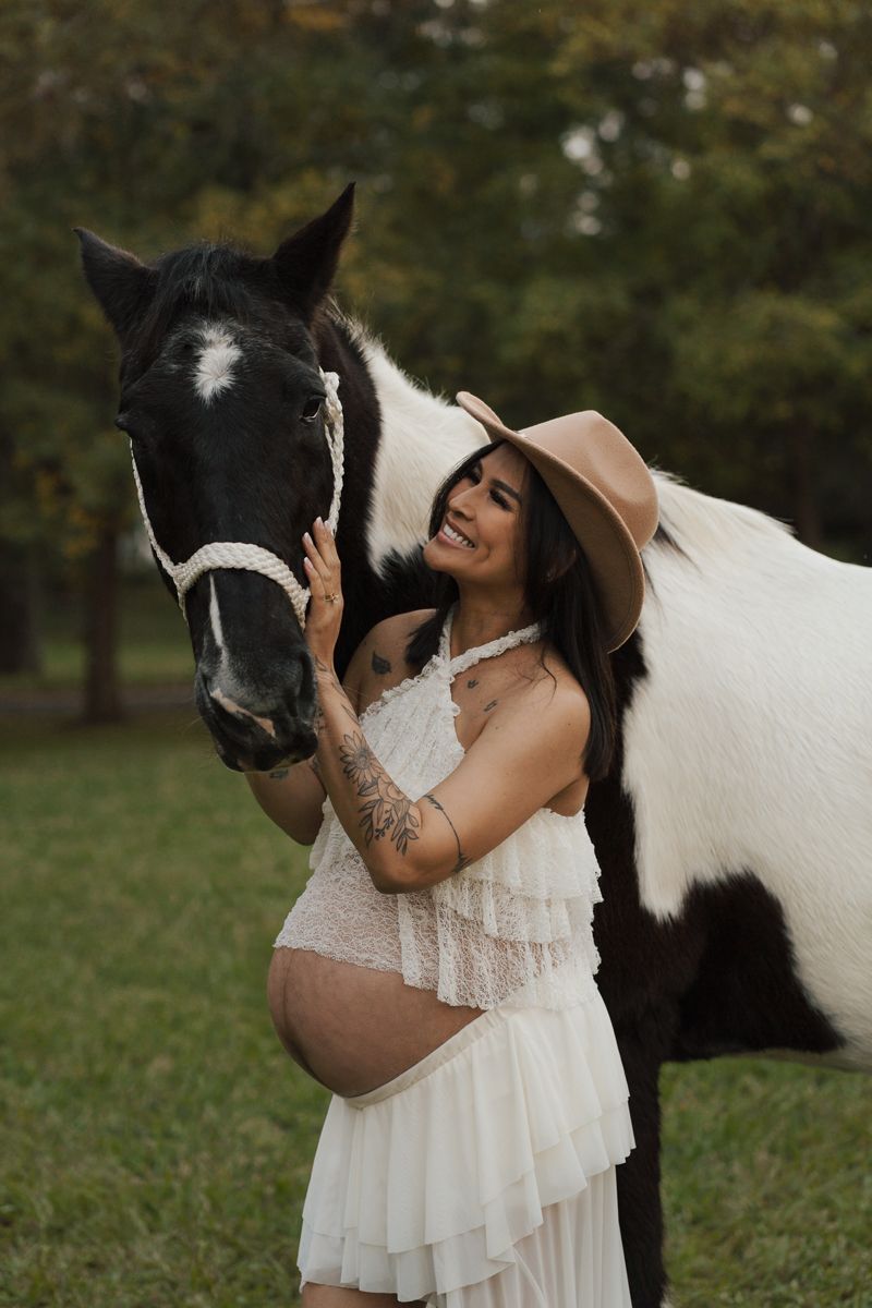 pregnant woman in white dress and cowboy hat poses with a black and white paint horse outdoors. maternity photoshoot with a horse in Sydney