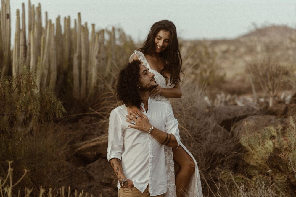 Couple in white outfits embracing among tall cacti in a desert landscape during golden hour.