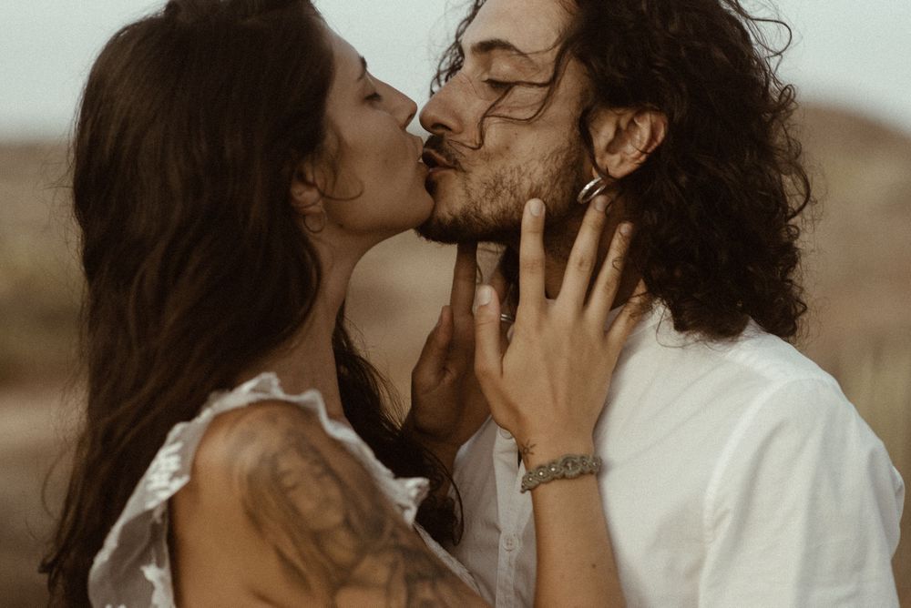 A couple shares a romantic kiss outdoors, the woman with tattoos gently holding the bearded mans face.