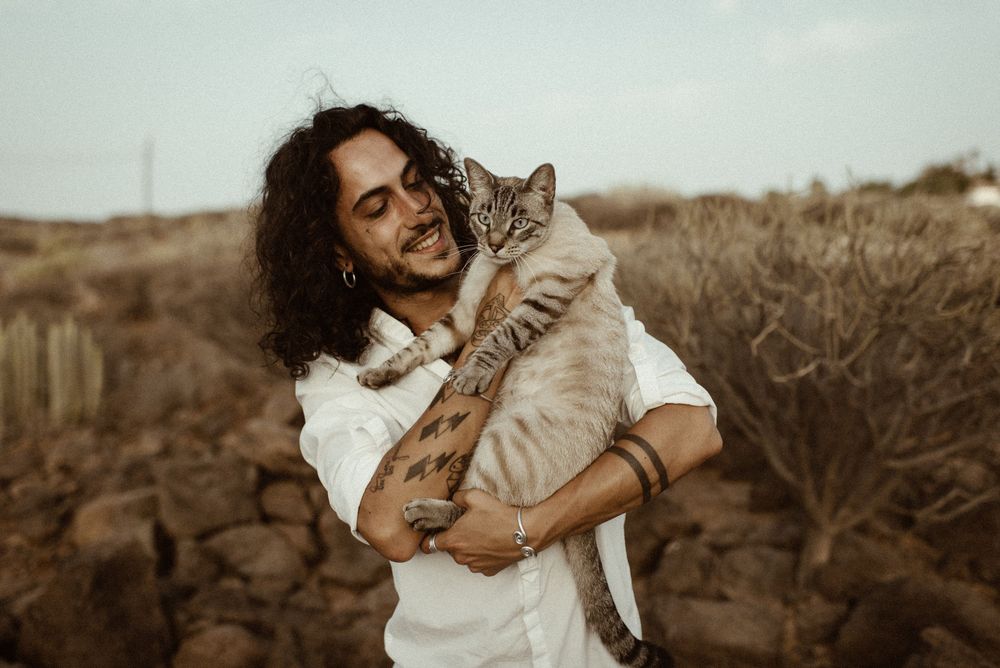A smiling man with long curly hair holds a tabby cat outdoors in a dry rocky landscape.