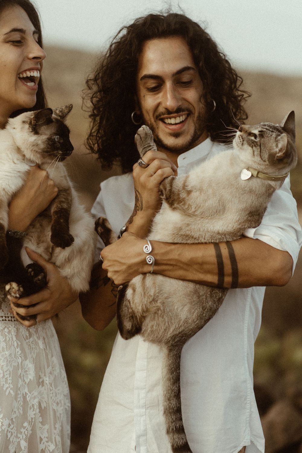 Smiling couple in white outfits holding two Siamese cats outdoors, laughing joyfully in a candid portrait.
