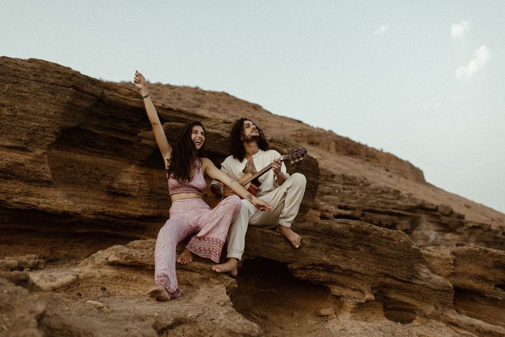 Two women sitting on desert rock formations, one pointing upward, wearing pink and white bohemian outfits.