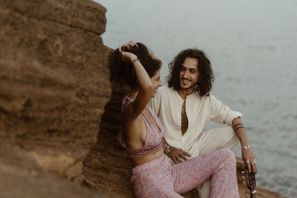 A smiling couple relaxes against a rocky cliff by the ocean, dressed in bohemian style outfits.