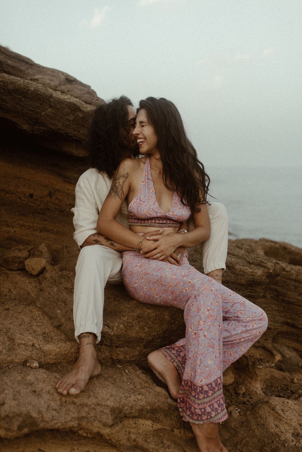 Couple sitting on coastal rocks, woman in pink boho outfit laughing as partner embraces her by the sea.