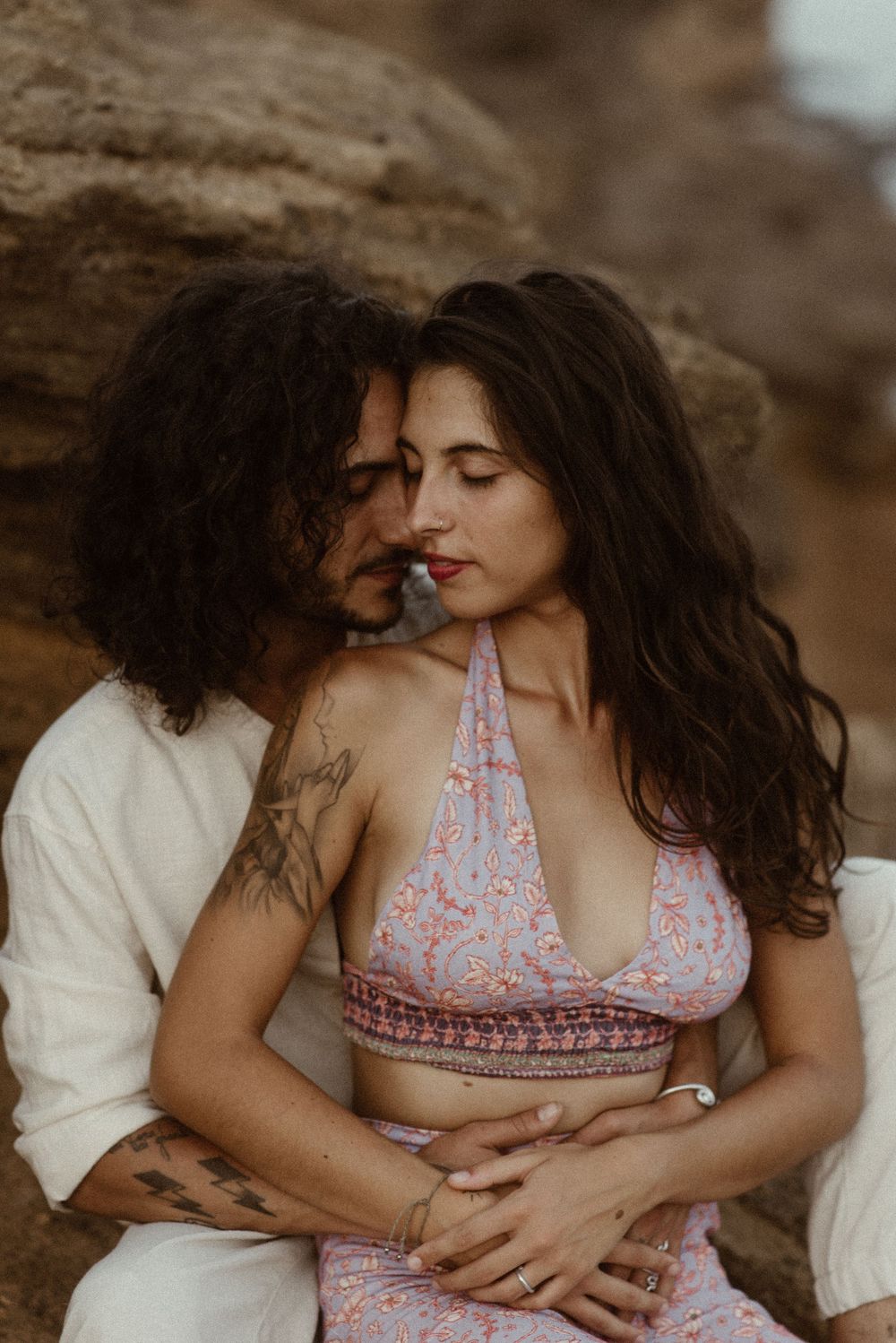 A tattooed couple shares an intimate moment, sitting together against rocky cliffs in a romantic outdoor setting.