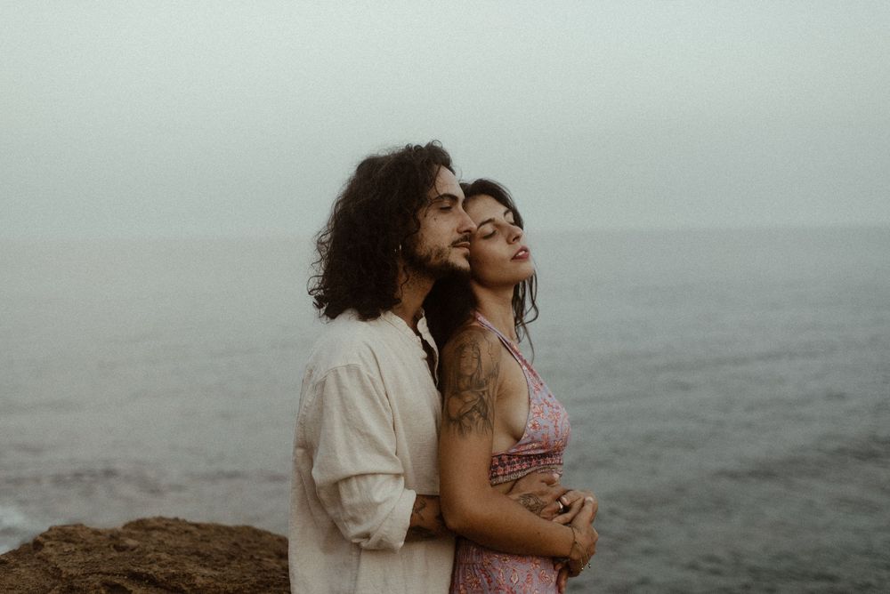 Romantic couple embracing by the ocean on a misty day, woman in floral dress with tattoo, moody coastal backdrop.