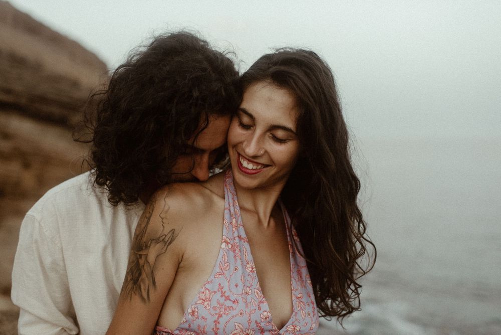 Happy couple sharing a tender moment by the sea, woman smiling joyfully as man nuzzles her cheek near rocky shore.
