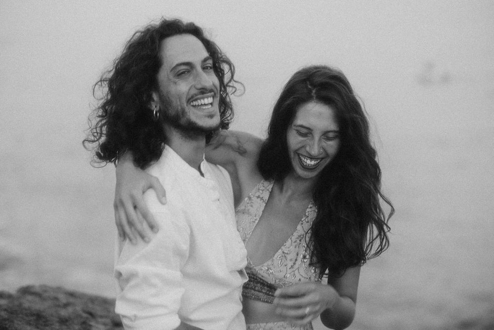 Black and white photo of a happy couple embracing and laughing together outdoors near a rocky shore.