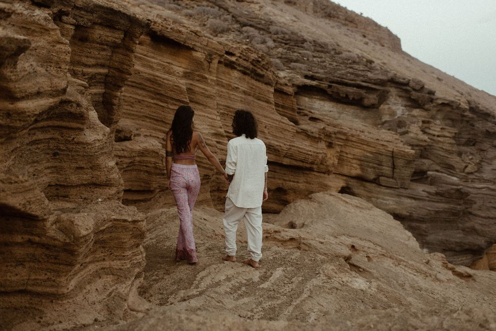 Two women standing before layered sandstone cliffs in pink and white outfits, viewed from behind.