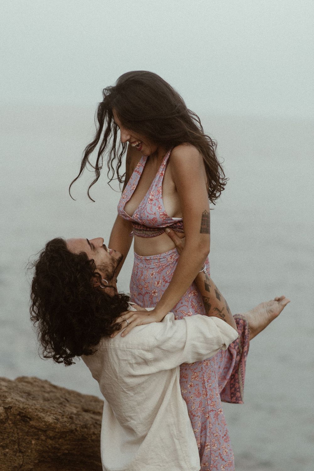 A man lifts a woman in a floral dress near the ocean, sharing a romantic moment on rocky shores.
