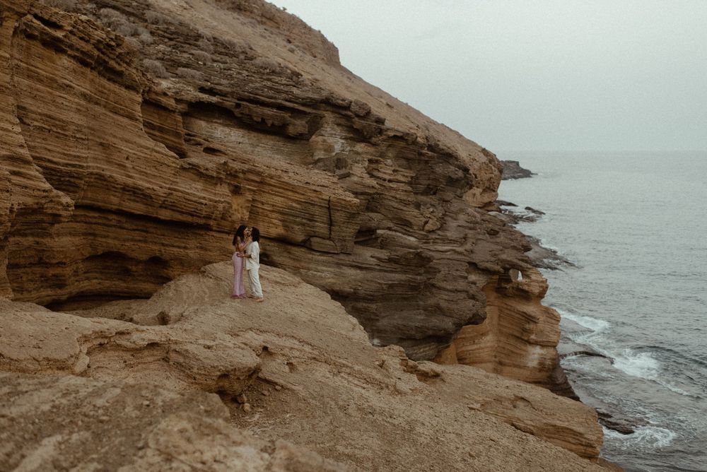Couple embracing on rugged coastal cliffs with layered sandstone rocks and calm ocean in the background.