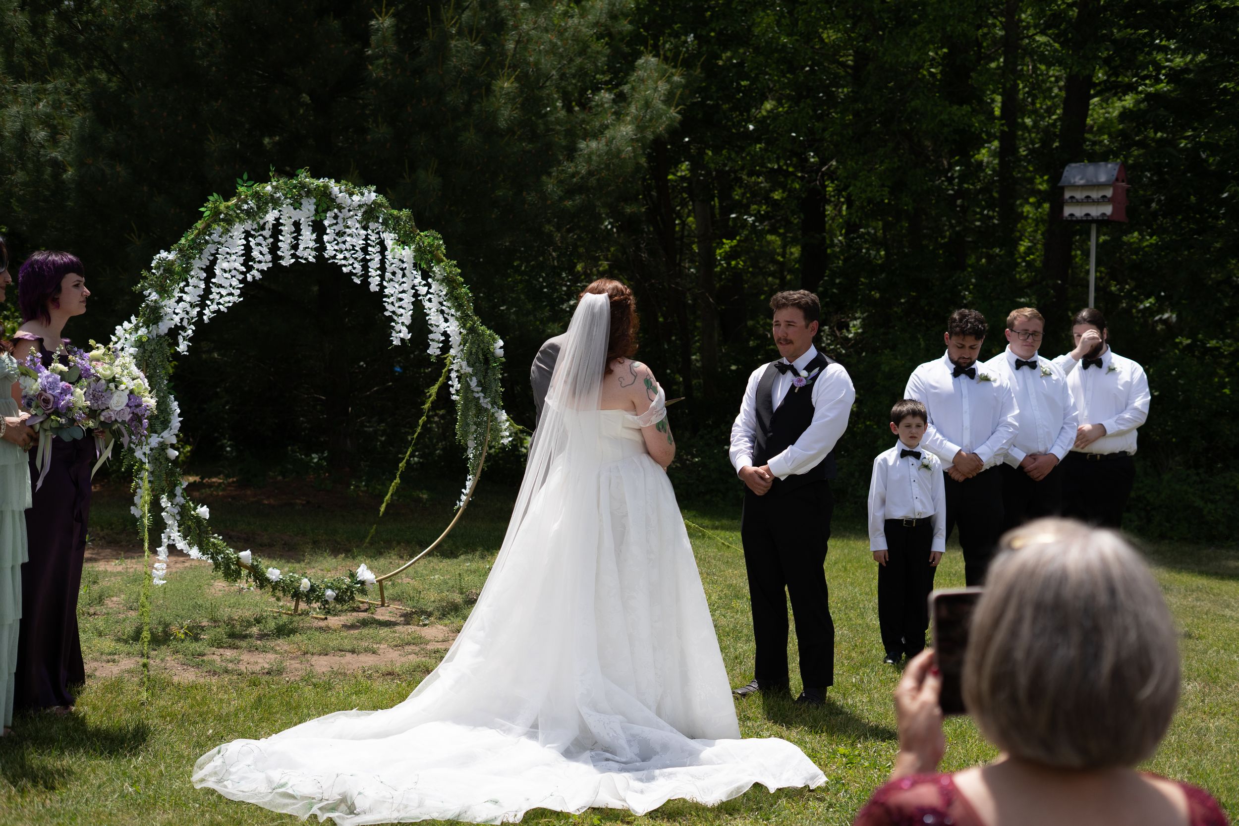 Bride and groom exchanging vows under a floral arch during an outdoor wedding ceremony with guests watching.