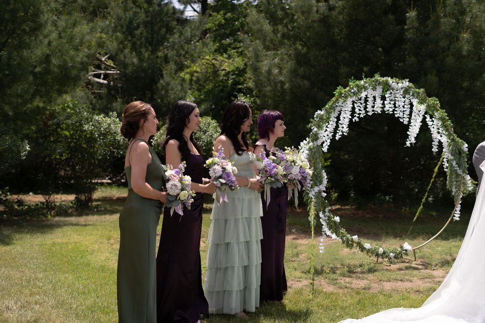 Bridesmaids in sage and purple gowns holding bouquets stand beside a floral arch at an outdoor wedding ceremony.