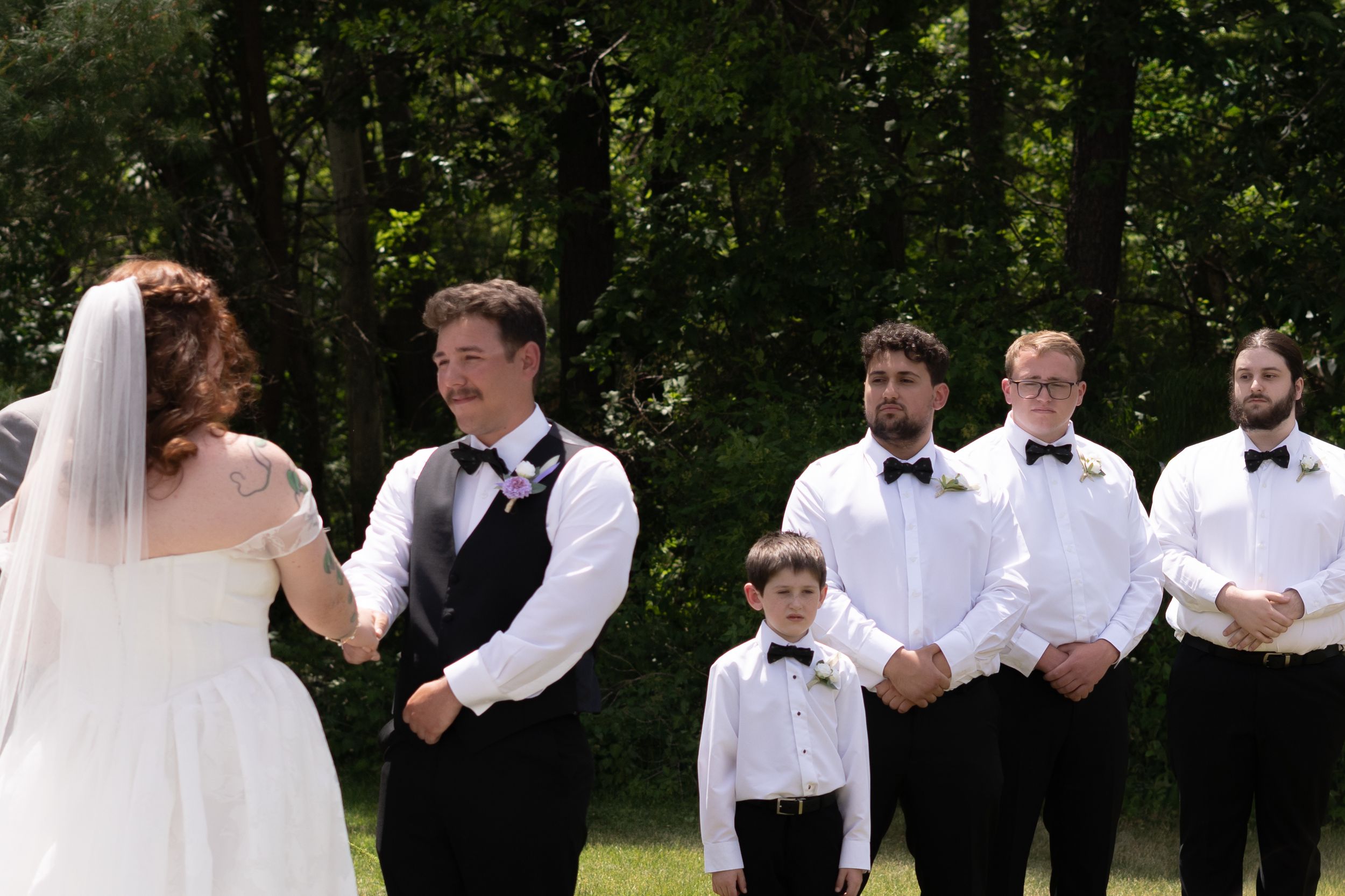 Groom with mustache holds bride's hands at altar with groomsmen and young ring bearer standing behind him outdoors.