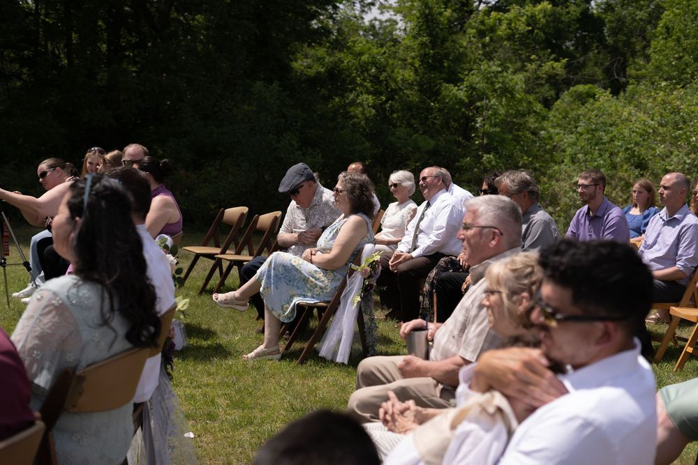 Wedding guests seated outdoors on sunny day watching ceremony, dressed in summer attire on green lawn.