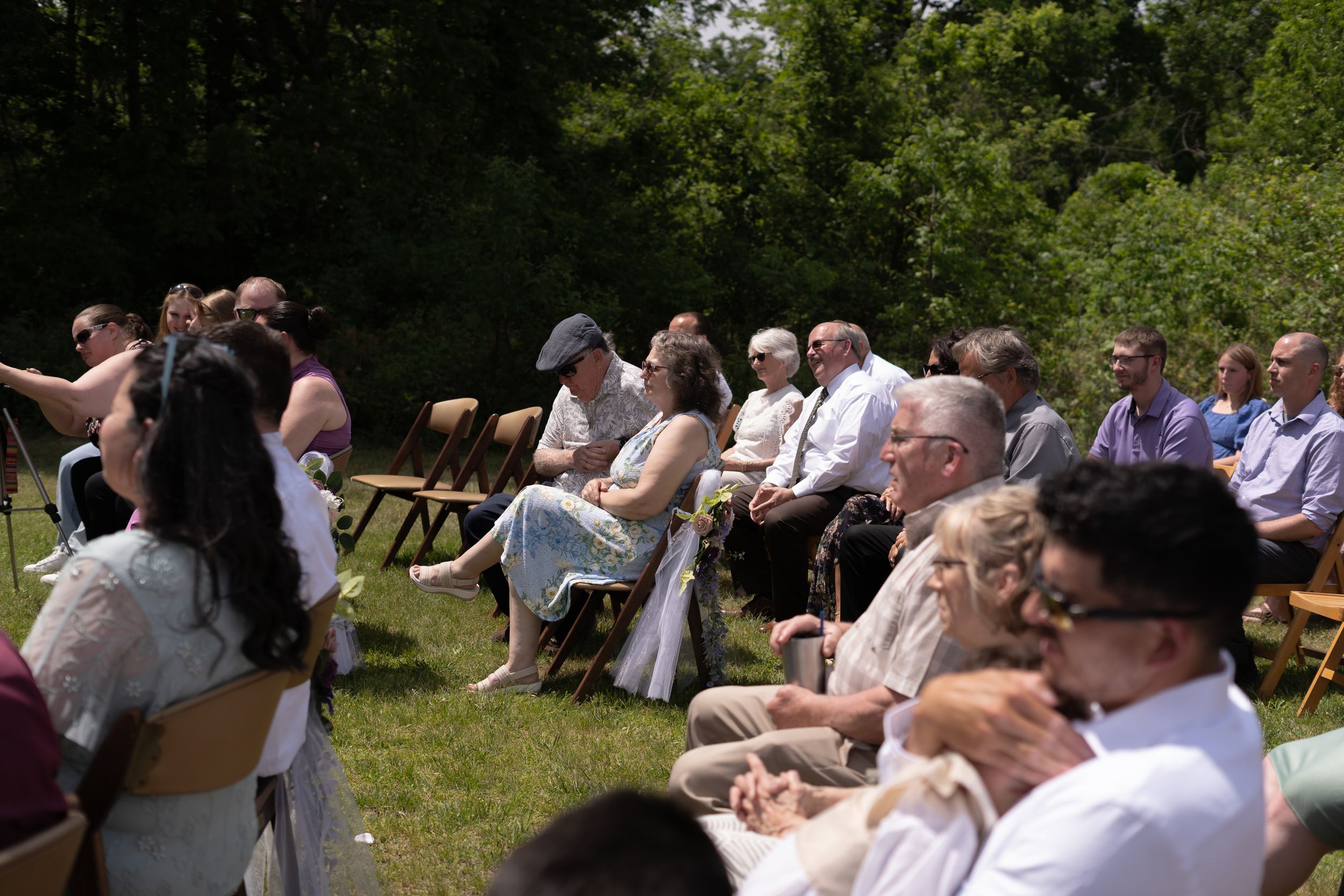 Wedding guests seated outdoors on sunny day watching ceremony, dressed in summer attire on green lawn.