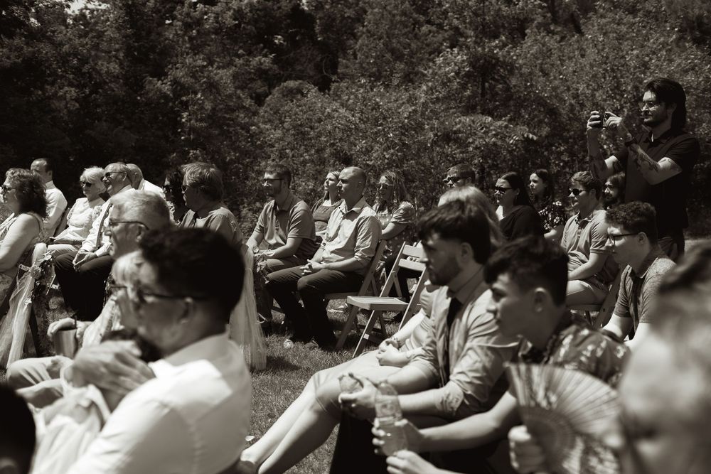 Black and white candid photo of wedding guests seated outdoors watching the ceremony in a garden setting.