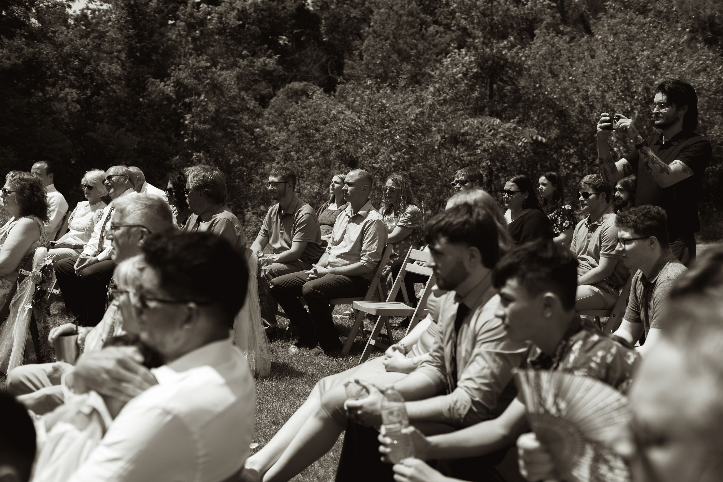 Black and white candid photo of wedding guests seated outdoors watching the ceremony in a garden setting.