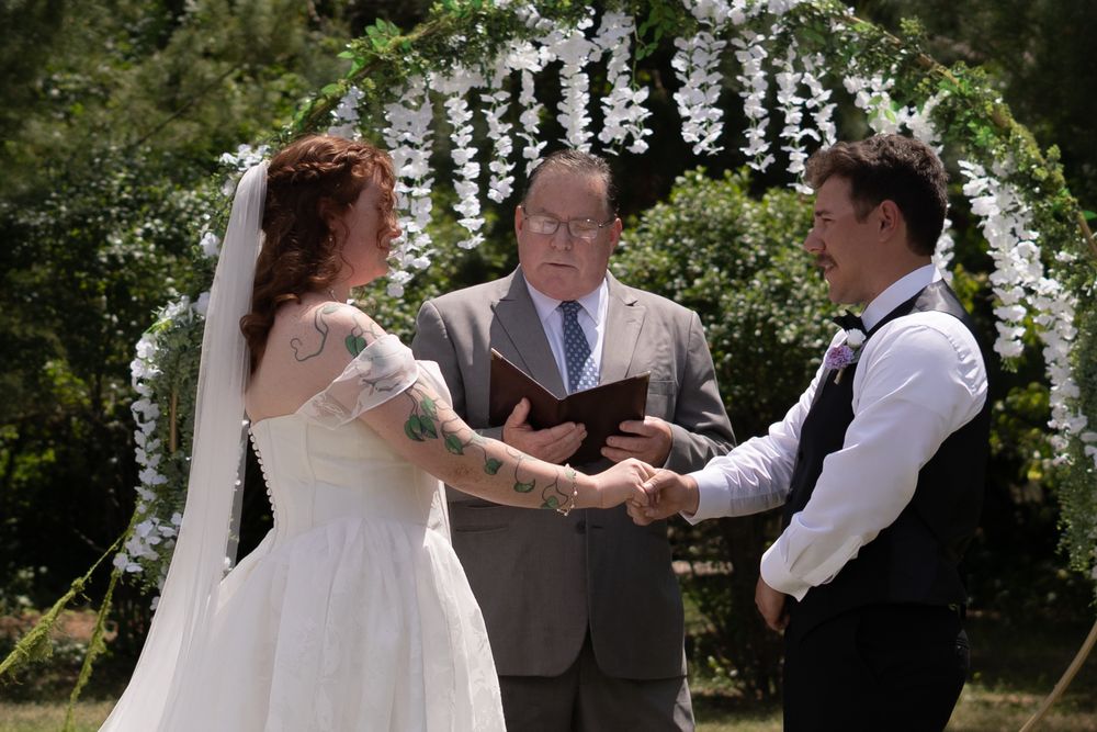 Bride and groom exchanging vows under floral arch with officiant at outdoor wedding.