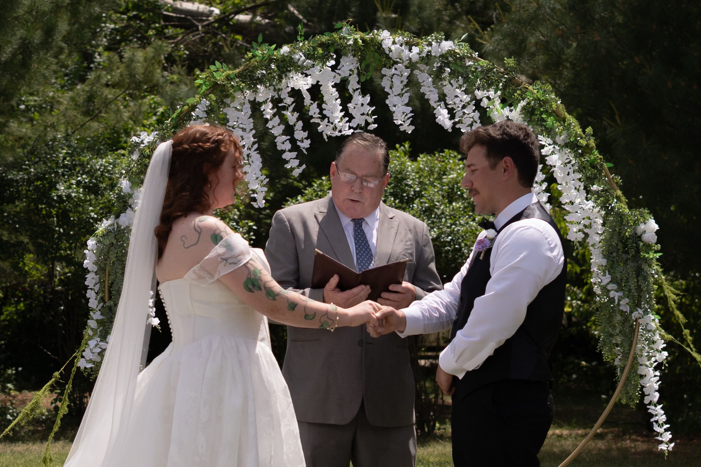Bride with tattoos and groom hold hands during outdoor wedding ceremony under white floral arch.