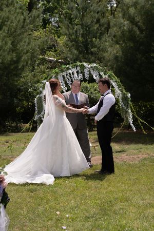 Bride in white gown and veil holding hands with groom at outdoor wedding altar adorned with white flowers.