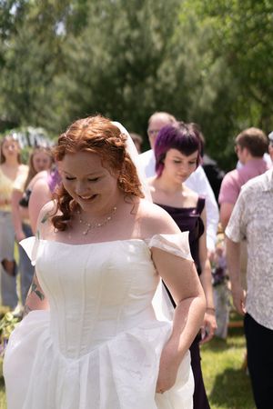 Smiling redhead bride in white off-shoulder gown walks outdoors surrounded by wedding party guests.