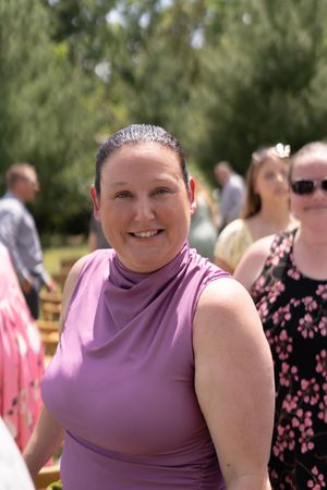 Smiling woman in purple sleeveless top poses at outdoor wedding celebration on sunny day.