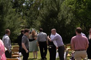 Wedding guests mingling and socializing outdoors near wooden chairs after garden ceremony.
