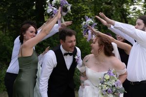 Wedding party celebrates with bride and groom raising purple bouquets in joyful outdoor ceremony.