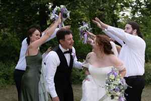 Wedding party celebrates outdoors raising bouquets with bride in white and bridesmaid in sage green.