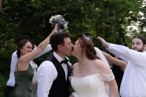 Groom kisses bride while wedding party raises bouquets overhead in lush outdoor garden setting.