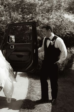Groom in black vest and bow tie holds open car door for bride in white wedding gown outdoors.