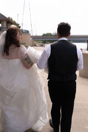 Bride and groom walk hand-in-hand from behind near waterfront venue after outdoor wedding ceremony.