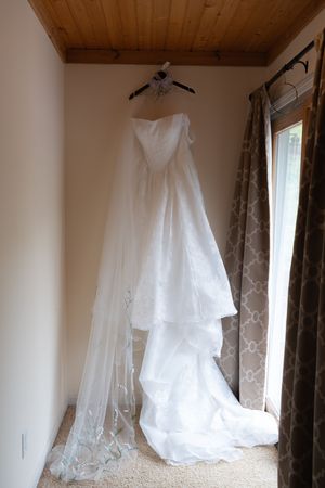 Elegant white wedding dress with lace veil hanging on hanger in room with wooden ceiling and window.
