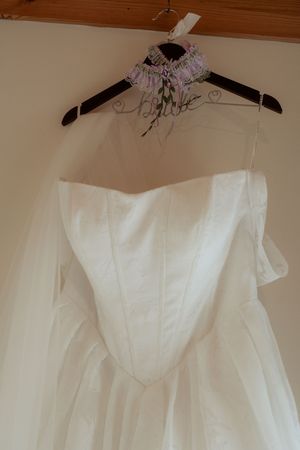 An off-shoulder white wedding gown hangs on a decorative wire hanger with a lavender garter on a cream wall.