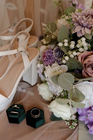 Bridal flat lay featuring white platform heels, purple floral bouquet, and wedding rings in green velvet boxes.