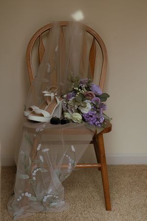 Wedding veil, white heels, and a purple floral bouquet displayed on a wooden chair.