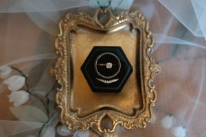 Diamond solitaire engagement ring displayed in dark hexagonal ring box on ornate gold decorative frame.
