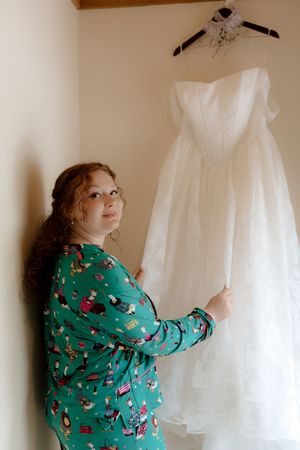 Bride in green pajamas holding white wedding dress on hanger in corner of room.