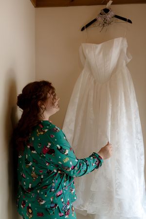 Bride in green pajamas admires her white strapless wedding gown hanging in a warmly lit room.