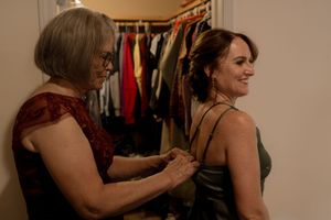 Older woman helps a smiling young woman fasten her dress in a closet room.