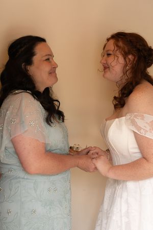 Bride in white gown holds hands with woman in blue lace dress in tender pre-wedding moment.