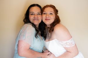 Bride in white off-shoulder gown embraces older woman in blue lace dress in warm indoor wedding photo.