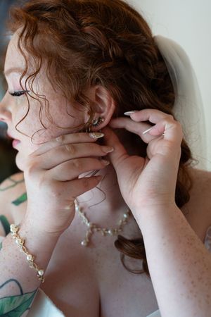 Close-up of redhead bride with freckles putting on earrings, wearing pearl necklace and bracelet.