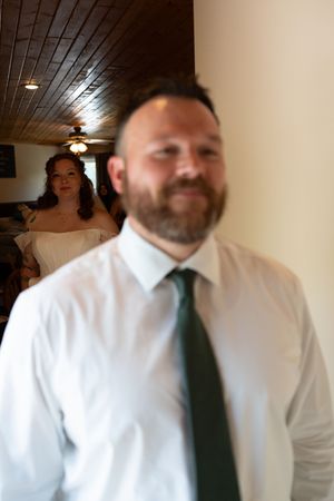 Bearded man in white shirt and green tie smiles indoors with bride visible in soft focus background.