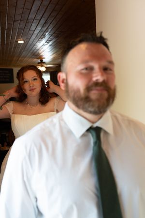 Red-haired bride in white gown poses behind bearded man in white shirt and green tie indoors.
