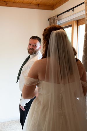 Bearded groom in suspenders smiles at bride in white veil and off-shoulder gown during first look.